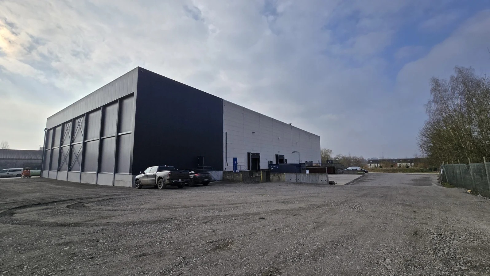 Zewnętrzny widok hali przemysłowej z czarno-białą elewacją, z rampami załadunkowymi i zaparkowanymi pojazdami na żwirowym placu, pod pochmurnym niebem.