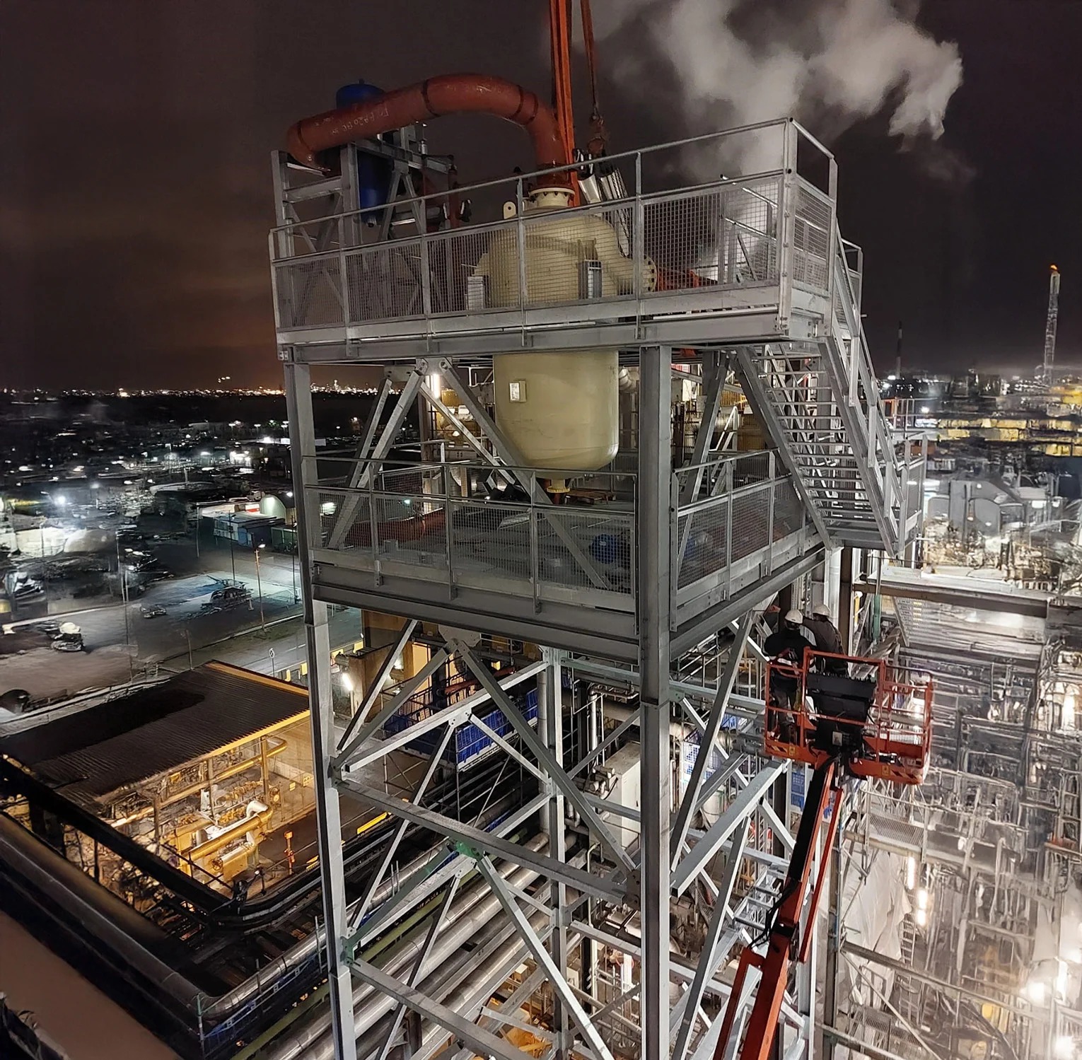 Nocne ujęcie stalowej konstrukcji wsporczej zbiornika impulsowego na terenie rafinerii, z widocznymi elementami instalacji i pracownikami na podnośniku.