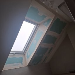 Jan Eisermann - Poddasze z oknem dachowym w trakcie prac wykończeniowych. Widoczne płyty gipsowo-kartonowe i szpachlowane łączenia. Oświetlenie sufitowe punktowe.