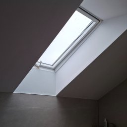 Jan Eisermann - Okno dachowe z roletą w łazience w Poznaniu. Szare płytki na ścianach, minimalistyczny design. Ujęcie z dołu, widoczny fragment półki z akcesoriami.