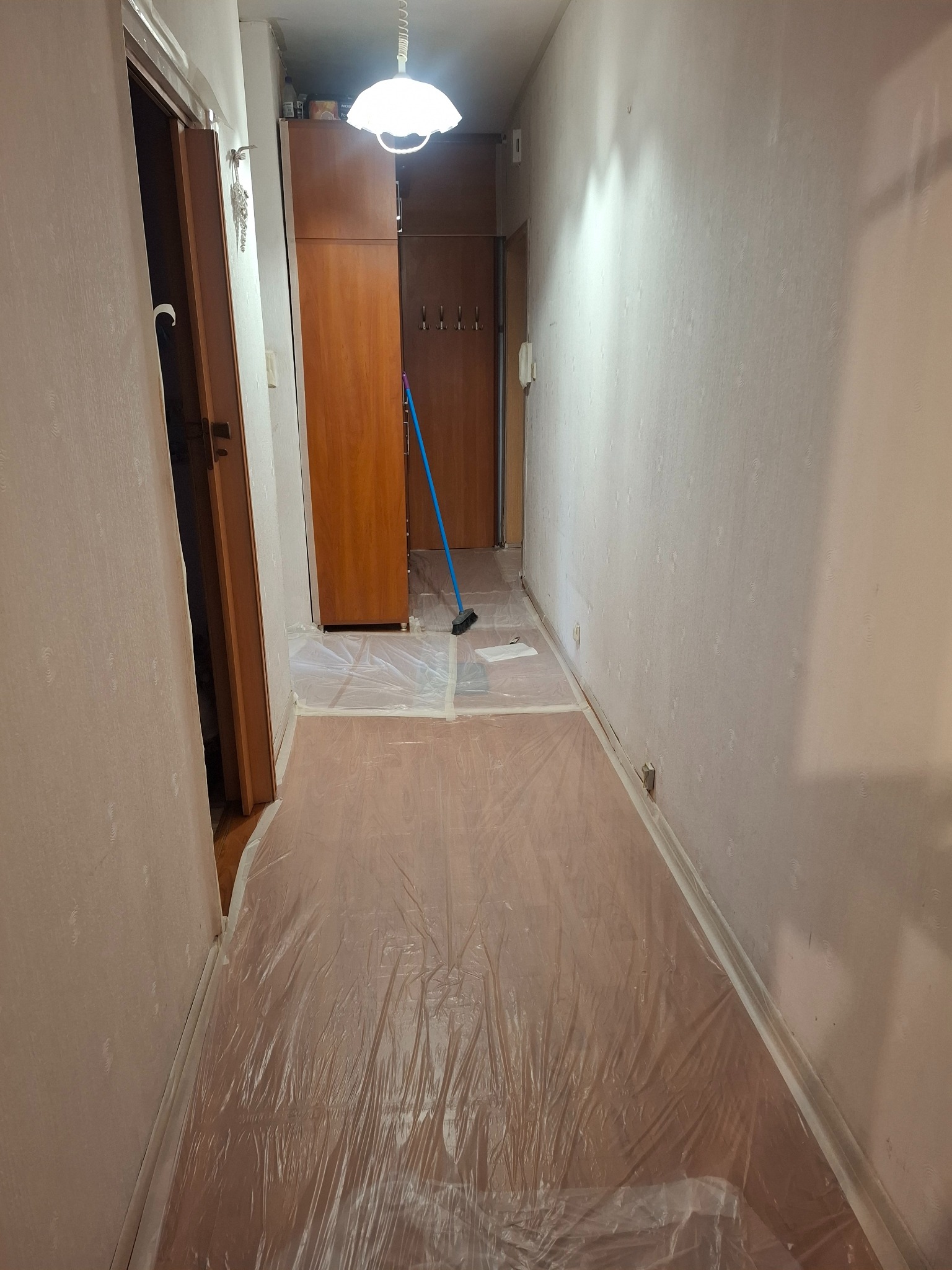 Korytarz zabezpieczony folią malarską przed remontem. Widoczne ściany z tapetą, drewniane drzwi i podłoga. Przy ścianie stoi miotła.
