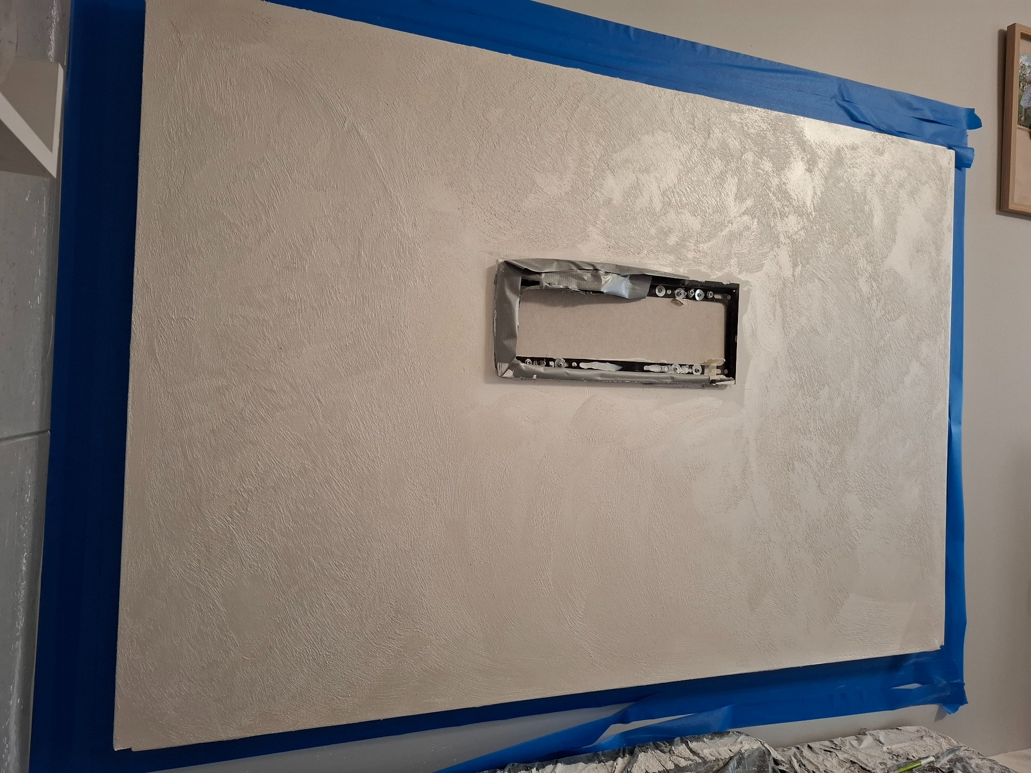 Ściana po szpachlowaniu dekoracyjnym, zabezpieczona niebieską taśmą malarską. Widoczny otwór na gniazdko zabezpieczony taśmą. Delikatna faktura na powierzchni ściany.