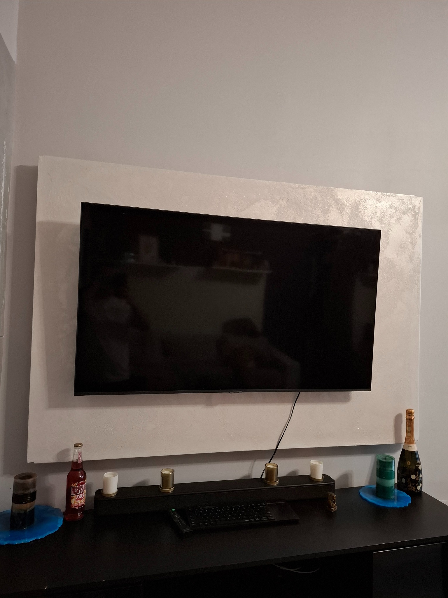 Telewizor na ścianie z dekoracyjną ramą o fakturze trawertynu, umieszczony nad czarną komodą z dekoracjami. Nowoczesny design wnętrza.