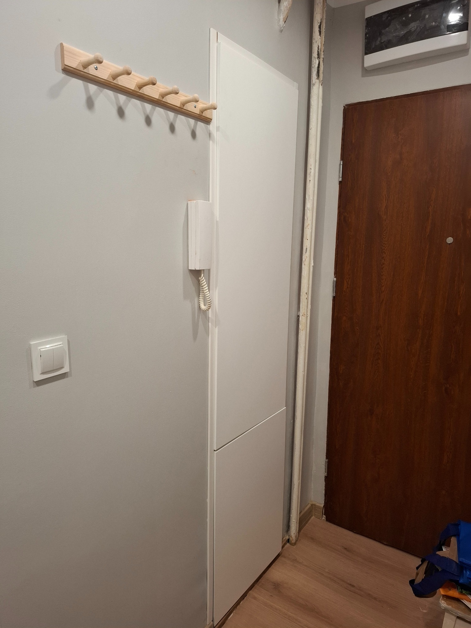 Szara ściana z drewnianym wieszakiem, białą szafą z domofonem i brązowymi drzwiami wejściowymi. Widoczny fragment rury grzewczej i włącznik światła.