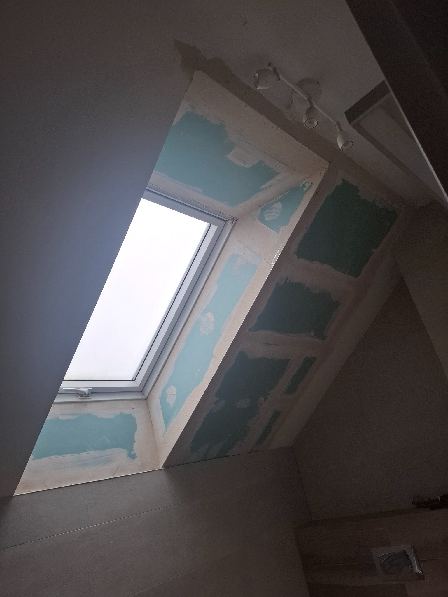 Poddasze z oknem dachowym w trakcie prac wykończeniowych. Widoczne płyty gipsowo-kartonowe i szpachlowane łączenia. Oświetlenie sufitowe punktowe.