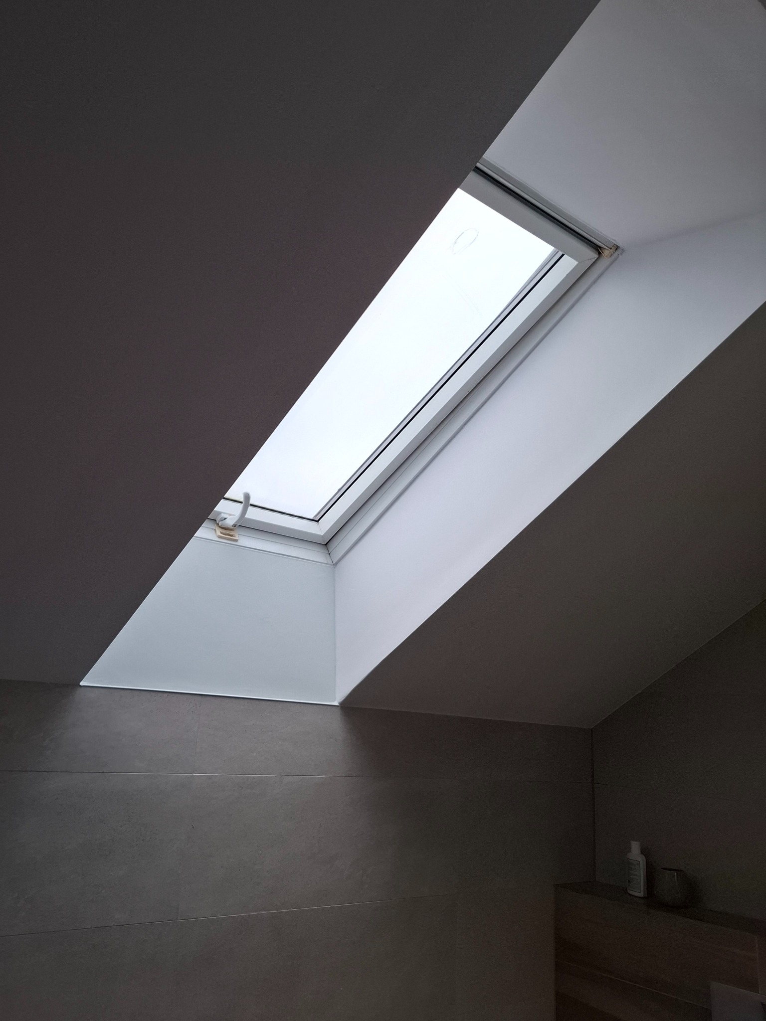 Okno dachowe z roletą w łazience w Poznaniu. Szare płytki na ścianach, minimalistyczny design. Ujęcie z dołu, widoczny fragment półki z akcesoriami.