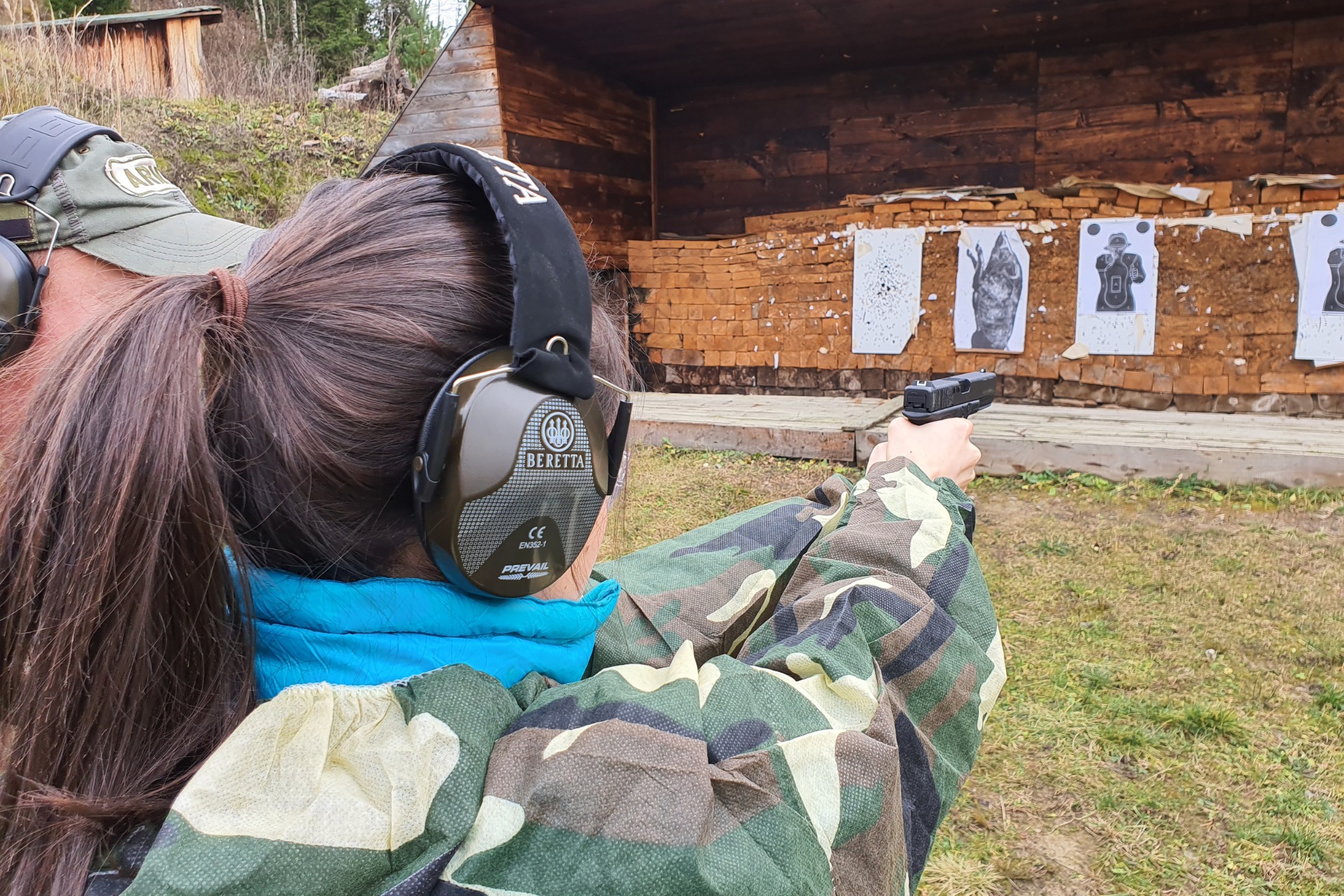 Strzelnica na świeżym powietrzu: kobieta w słuchawkach Beretta celuje z pistoletu do tarcz na drewnianej ścianie. Widok zza pleców, kamuflaż.