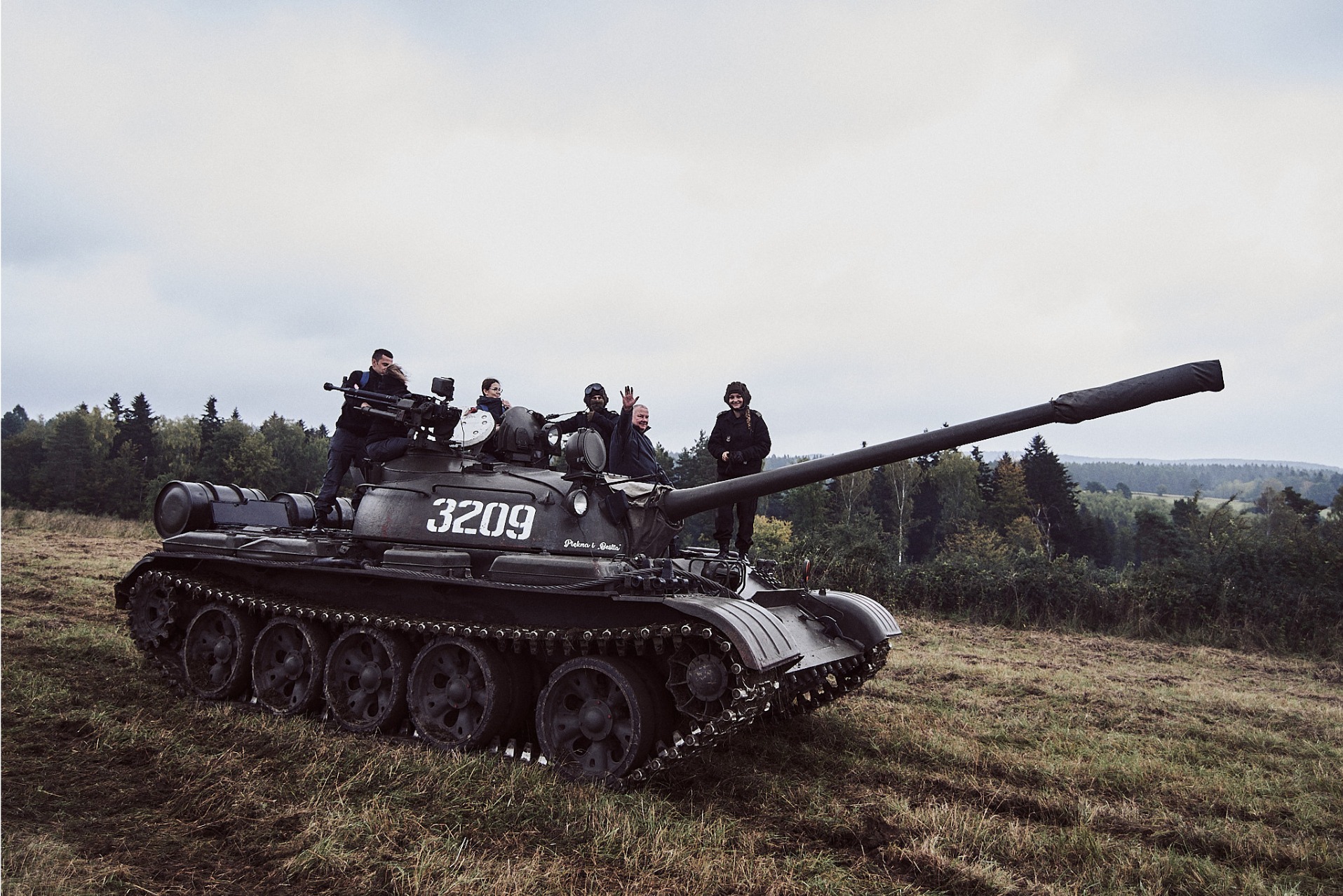 Czarny czołg T-55 z numerem 3209 na tle łąki i lasu. Na czołgu stoi grupa osób, w tym jedna machająca. Nietypowa atrakcja na imprezę plenerową.