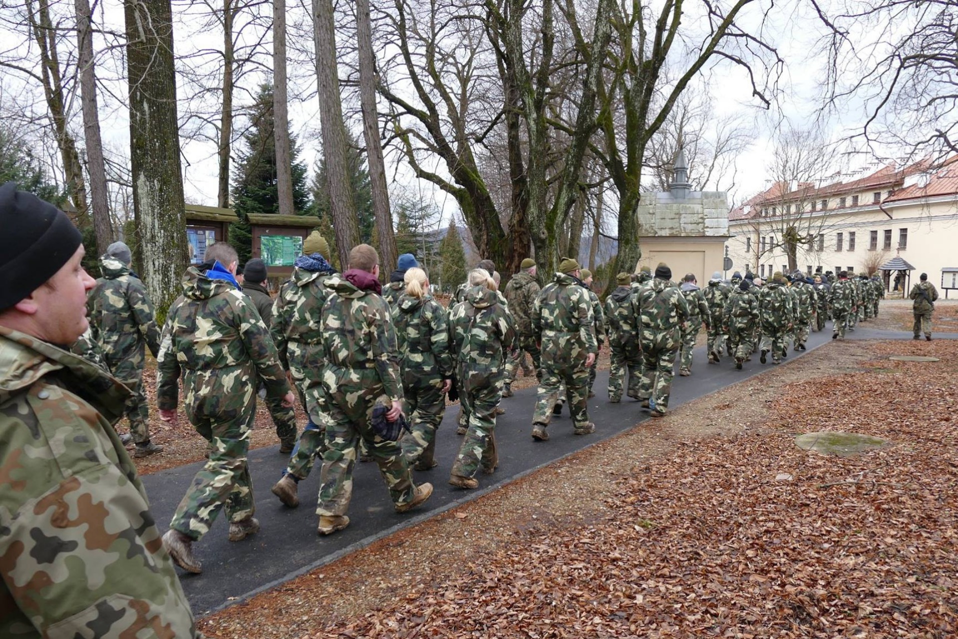 Grupa osób w strojach moro maszeruje aleją w parku, prawdopodobnie podczas imprezy integracyjnej o tematyce militarnej. W tle budynek i drzewa.