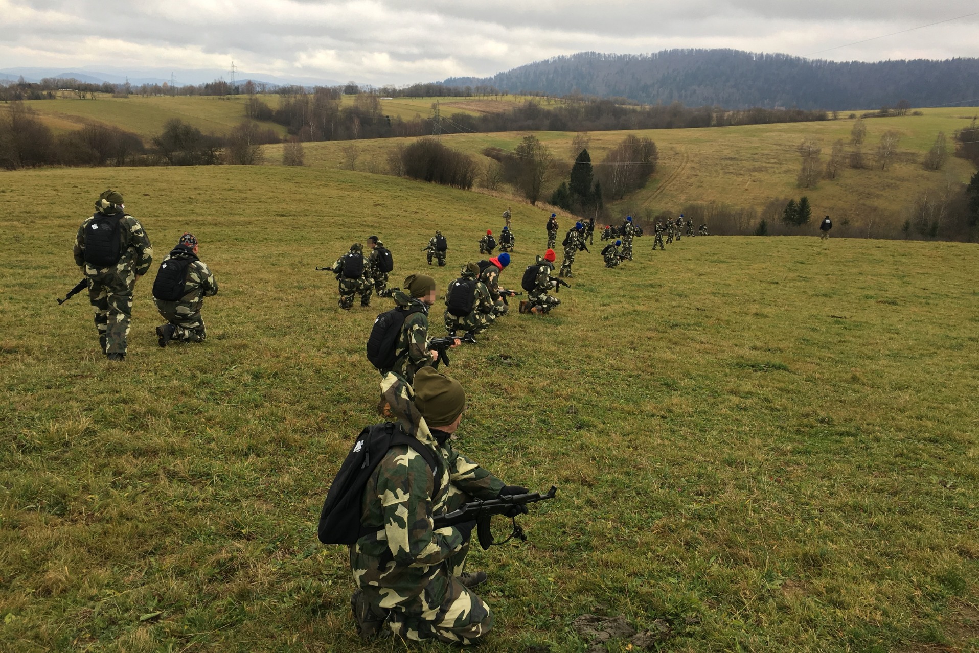 Grupa osób w strojach moro, z atrapami broni, na zielonym wzgórzu w Kwaszeninie, podczas gry terenowej. W tle las i pagórkowaty krajobraz.