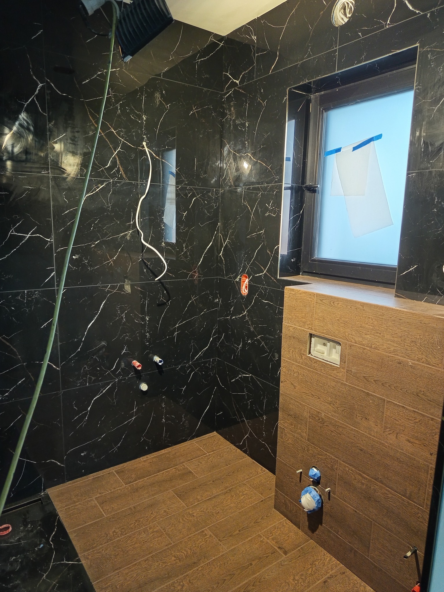 Ciemna łazienka w trakcie wykańczania: marmurowe płytki na ścianach, drewnopodobna zabudowa WC, okno z zabezpieczeniem. Widoczne instalacje przed montażem armatury.