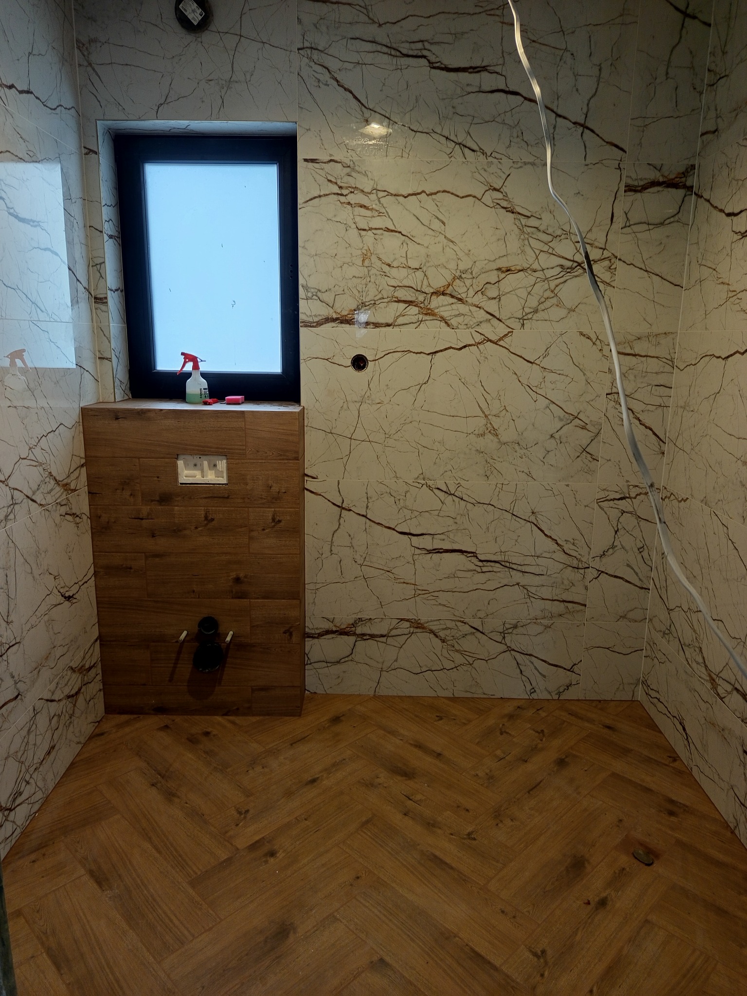 Wykończona łazienka z marmurowymi płytkami, drewnianą podłogą w jodełkę i zabudową WC. Widoczne okno i akcesoria łazienkowe. Nowoczesny design.