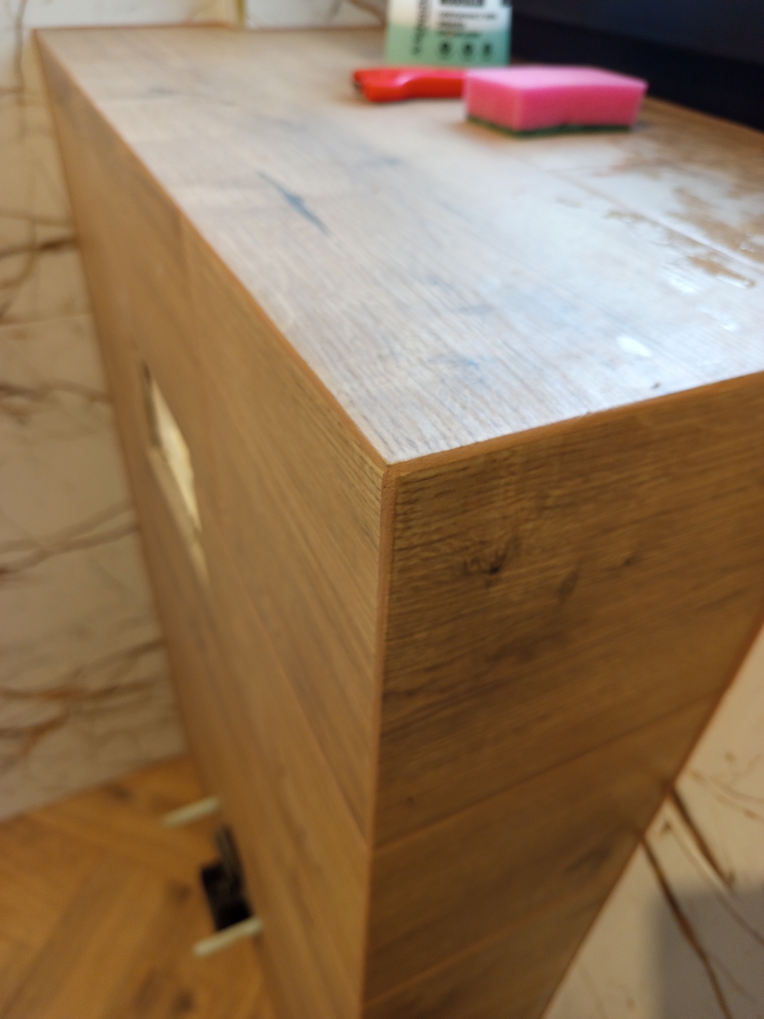 Drewniana szafka łazienkowa z blatem, na którym leży różowa gąbka i czerwona szpatułka. Widoczne płytki na ścianie i podłodze. Ujęcie z bliska, z góry.