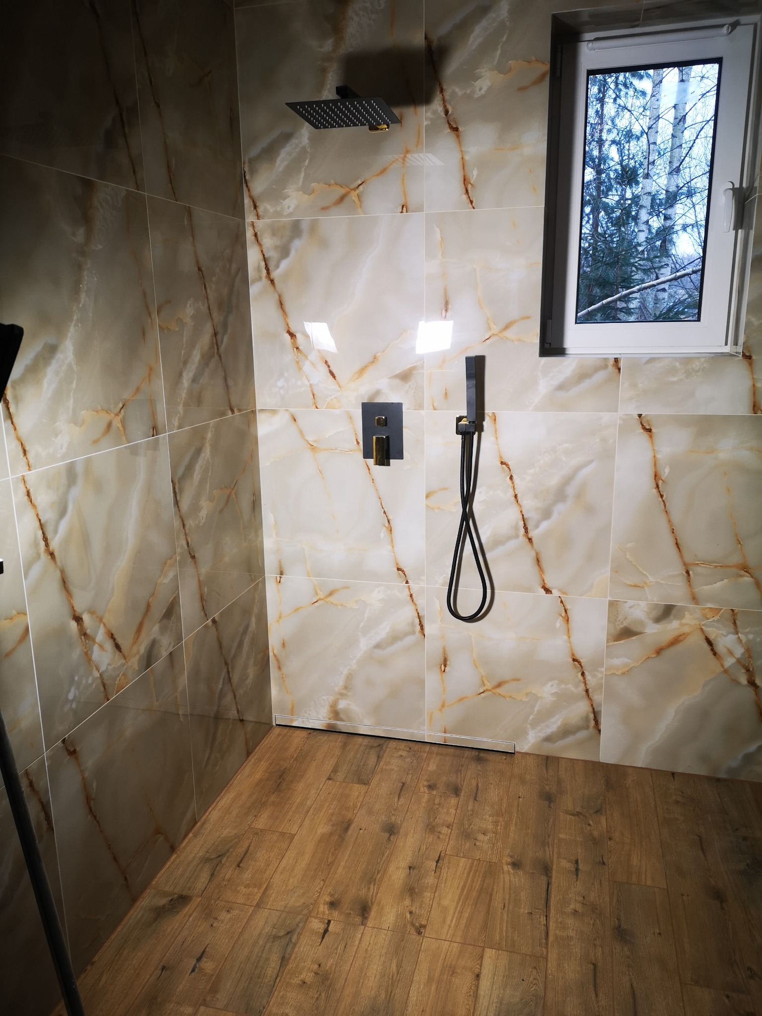 Wykończona łazienka z beżowymi płytkami imitującymi marmur, drewnopodobną podłogą i nowoczesną armaturą w kolorze czarnym. Widok na okno z widokiem na drzewa.