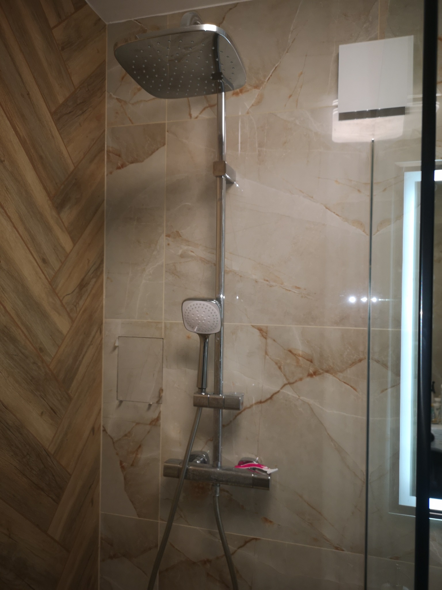 Nowoczesna kabina prysznicowa z deszczownicą i marmurowymi płytkami w odcieniach beżu i brązu. Widoczny fragment szklanej ściany i oświetlone lustro.