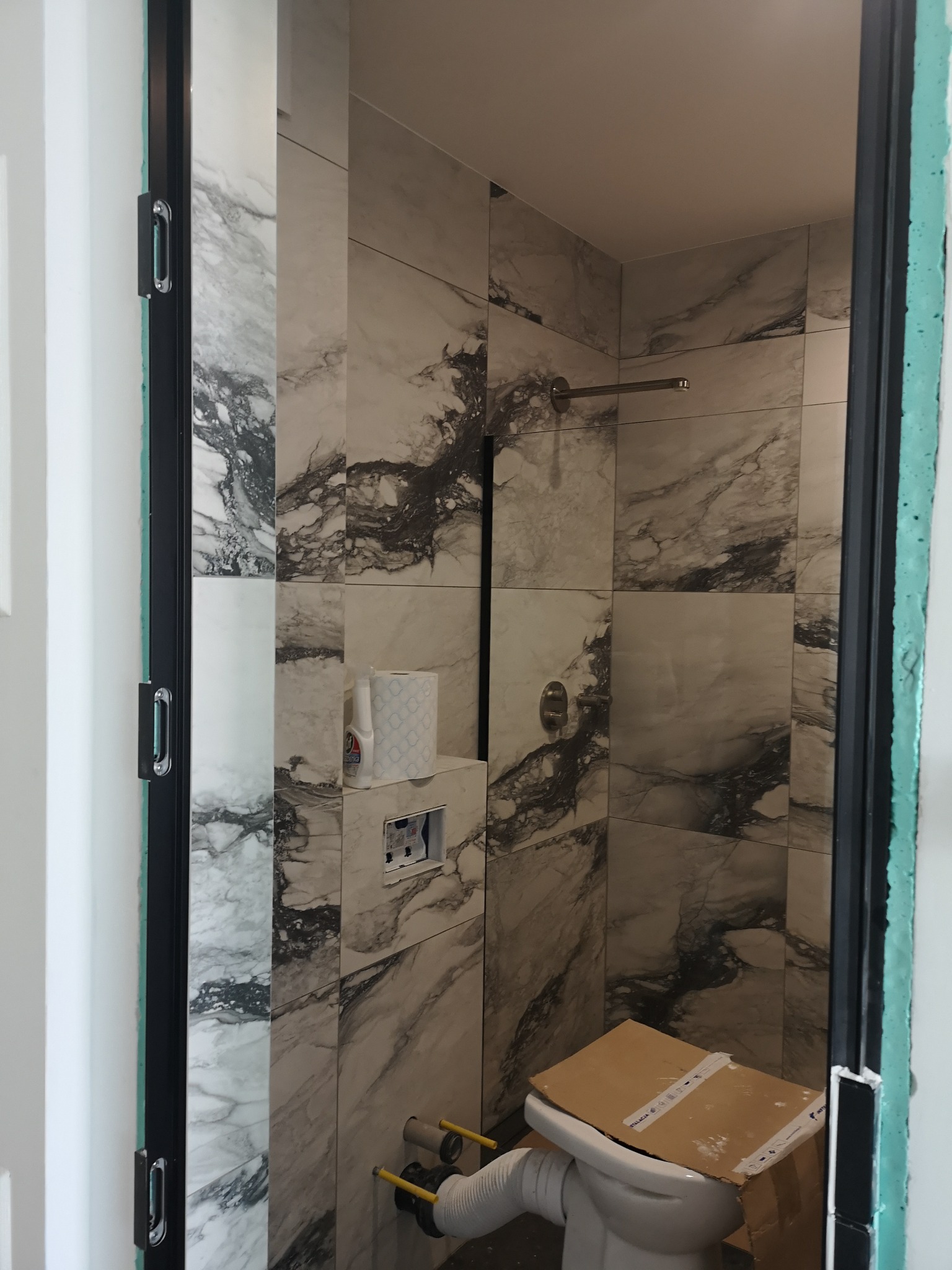 Wykończona łazienka z marmurowymi płytkami, widok przez futrynę drzwi. Częściowo zamontowana toaleta z kartonem na górze. Widoczne rury i elementy instalacji.