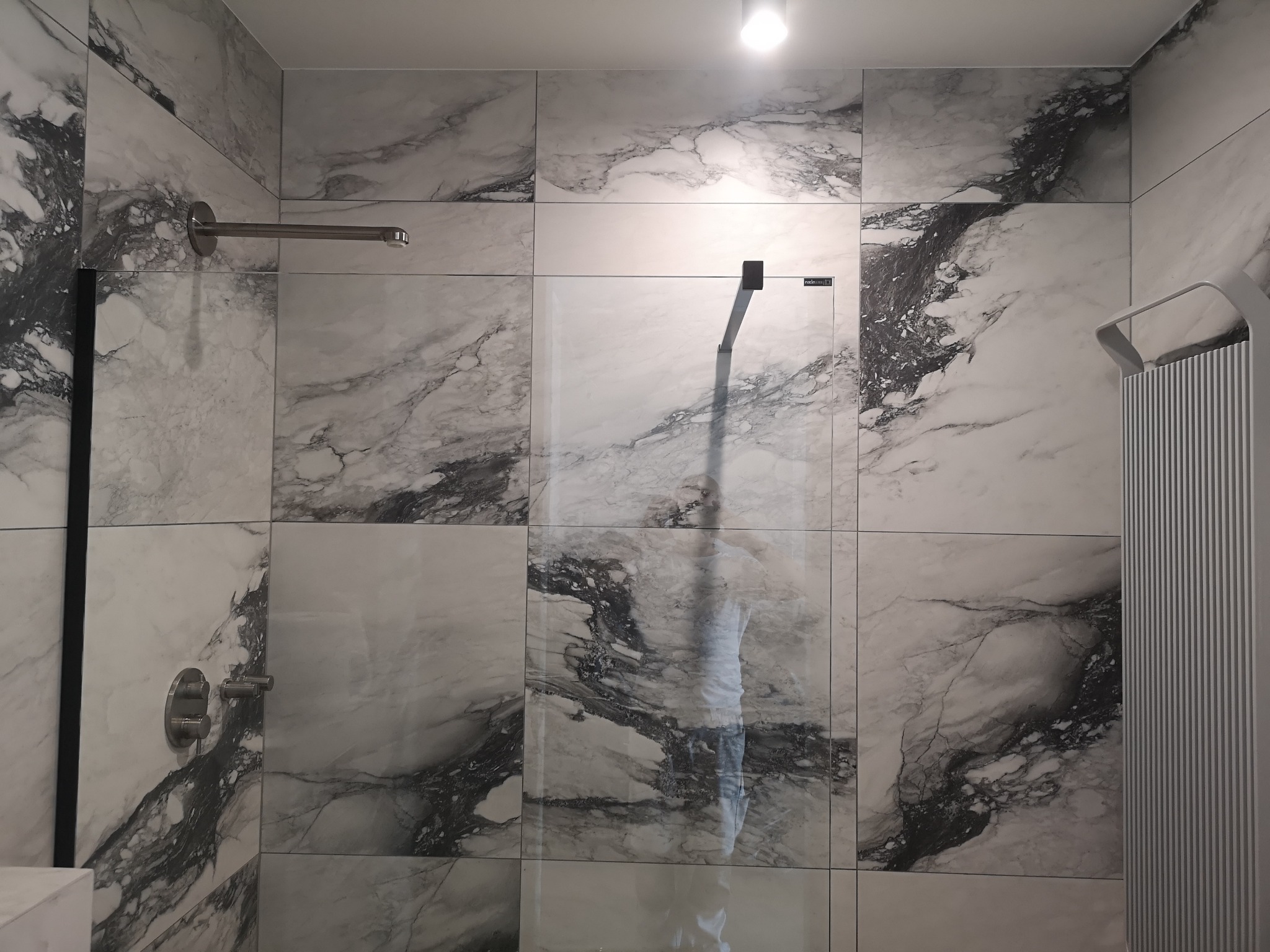 Wykończona kabina prysznicowa z marmurowymi płytkami, szklaną ścianką i grzejnikiem drabinkowym. Widoczne detale armatury i oświetlenie sufitowe.