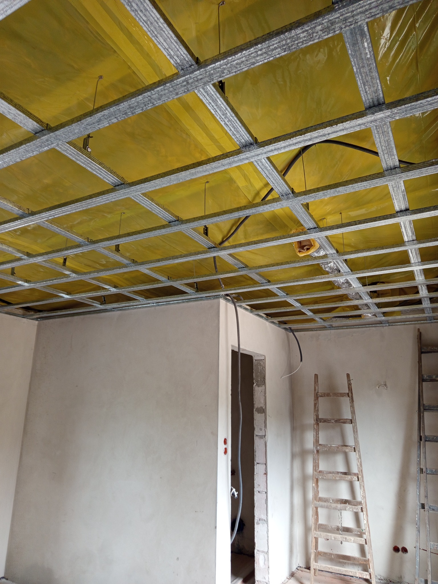 Montaż sufitu podwieszanego z metalowej konstrukcji i żółtej izolacji. Widoczne drabiny i ściany w trakcie prac wykończeniowych. Ujęcie z dołu.