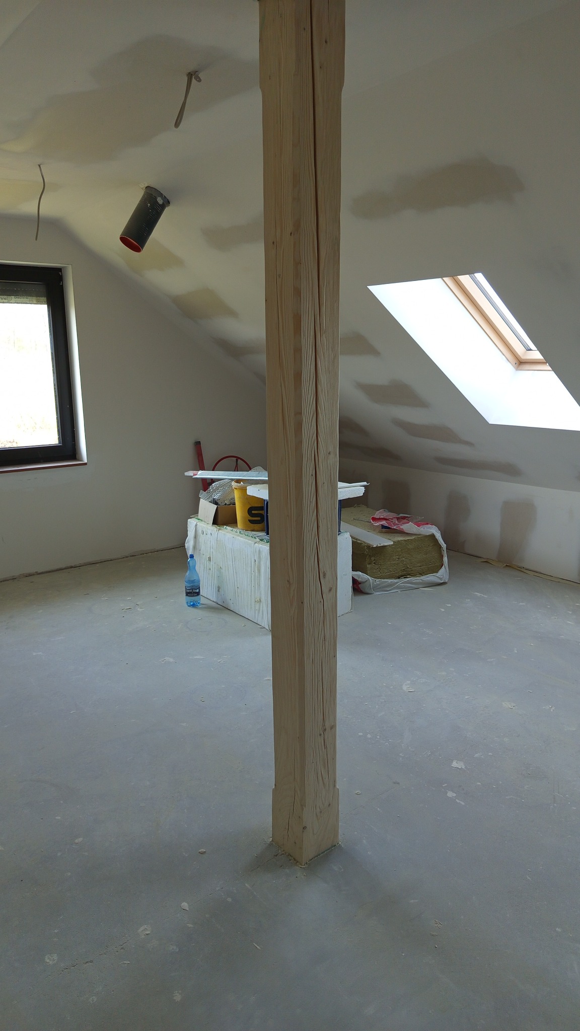 Poddasze w trakcie prac wykończeniowych: drewniany słup, okno dachowe, ślady szpachlowania na ścianach, materiały budowlane w tle. Widok na remontowany pokój.