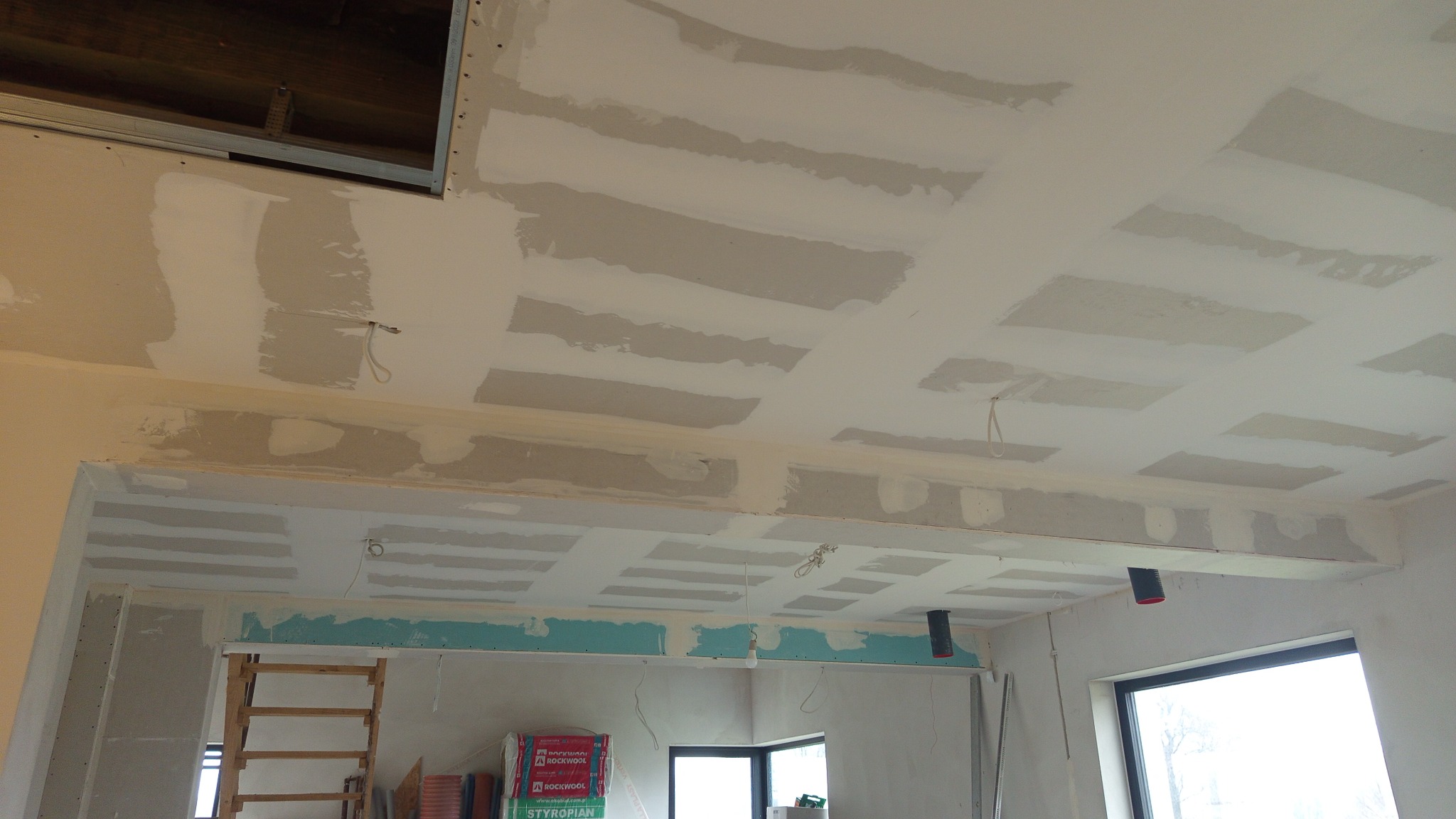 Sufit podwieszany z płyt gipsowo-kartonowych w trakcie szpachlowania, widoczne łączenia płyt i przewody elektryczne. W tle drabina i materiały budowlane.