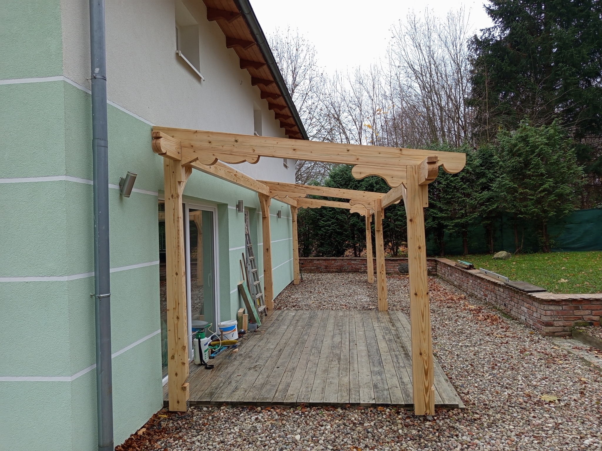 Drewniana pergola z dekoracyjnymi wycięciami, częściowo zmontowana przy domu z zieloną elewacją. Widoczne narzędzia i materiały budowlane. Jesienna sceneria.