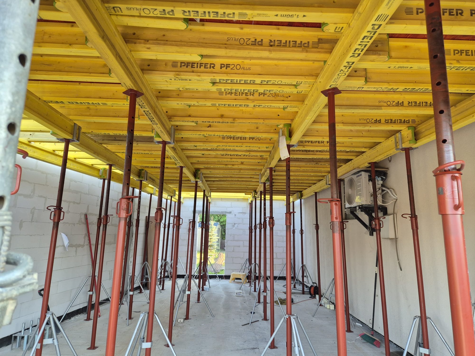 Żółte, drewniane szalunki stropowe wsparte na czerwonych, metalowych podporach w surowym wnętrzu budynku w trakcie budowy. Widoczne białe ściany z bloczków.