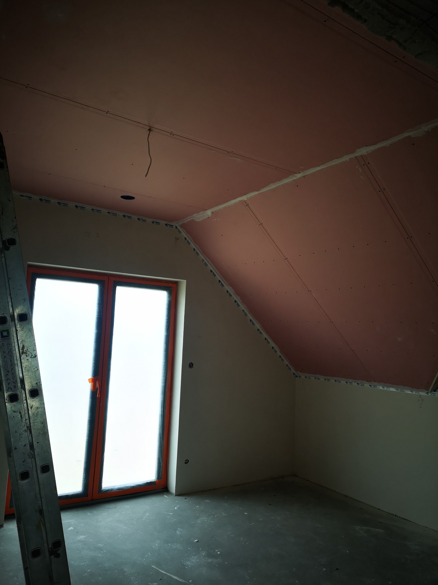 Poddasze w trakcie wykańczania: ściany i skośny sufit obłożone płytami gipsowo-kartonowymi, widoczna drabina oparta o ścianę, okno z pomarańczową ramą.