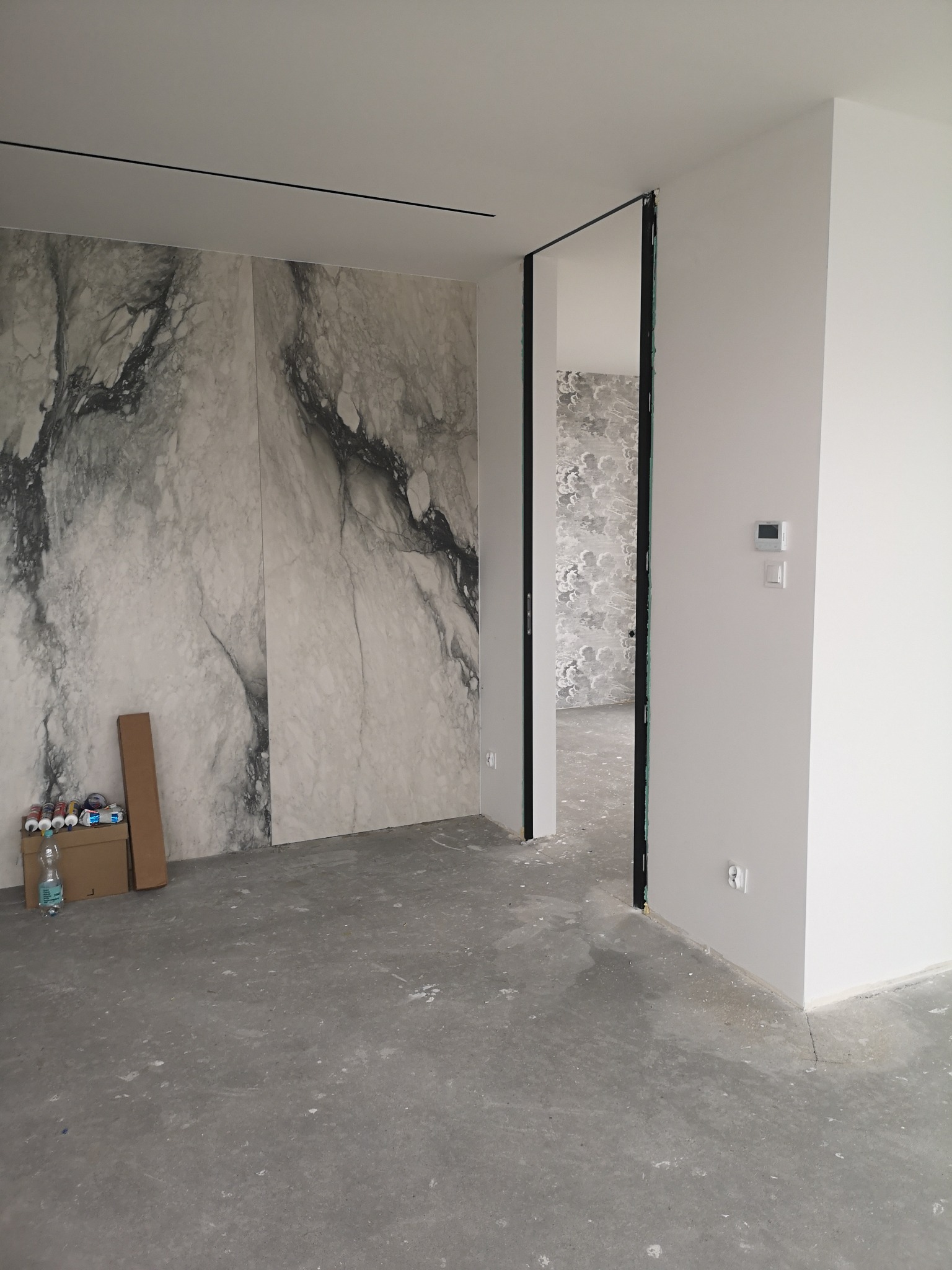 Wnętrze w trakcie wykańczania: ściana z efektem marmuru, białe ściany, surowa podłoga. Widoczne farby i narzędzia. Przejście do kolejnego pomieszczenia.