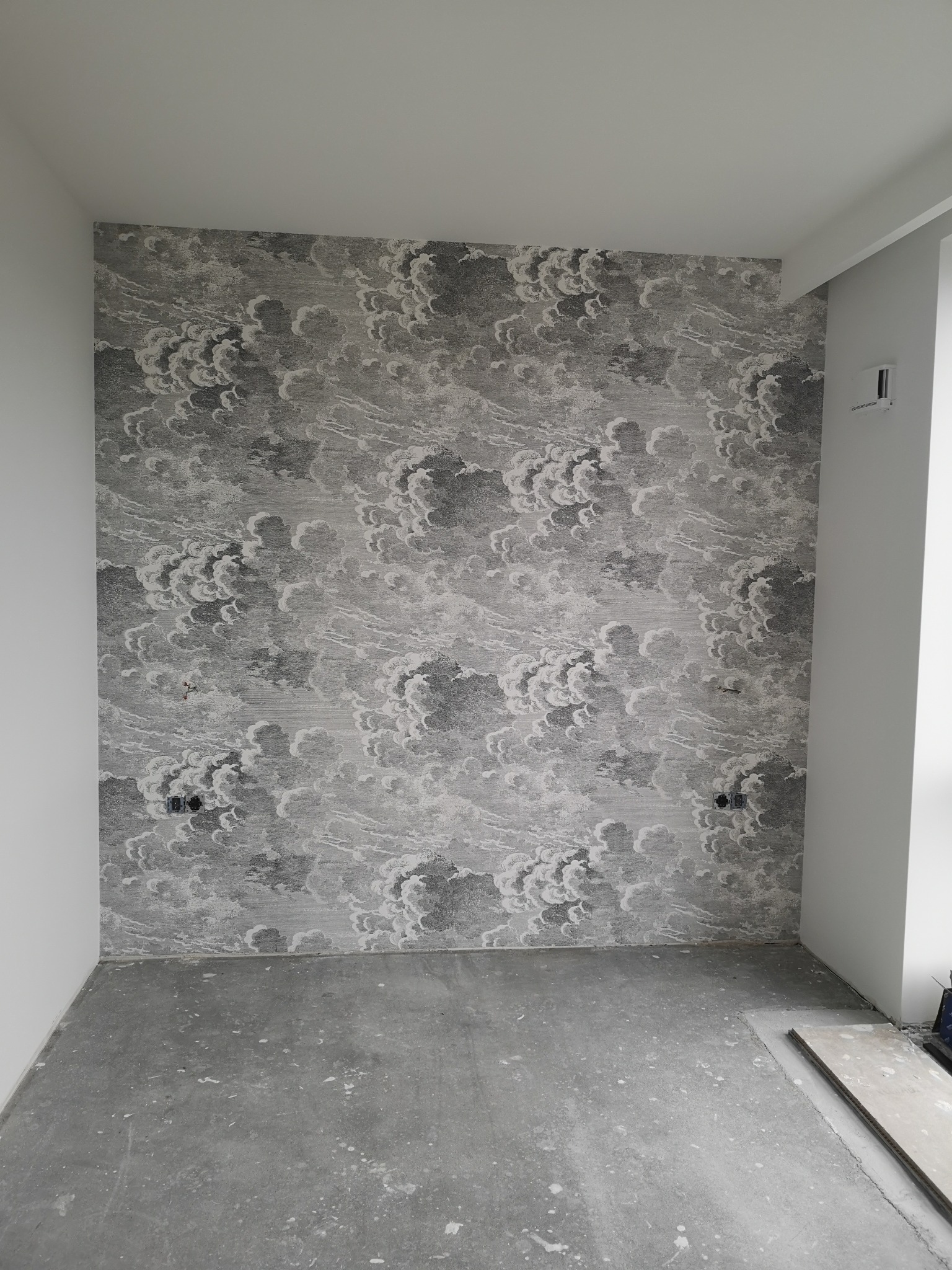 Pokój po tapetowaniu: ściana w szaro-białe wzory chmur, gładkie białe ściany boczne, surowa podłoga. Widoczne gniazdka elektryczne.