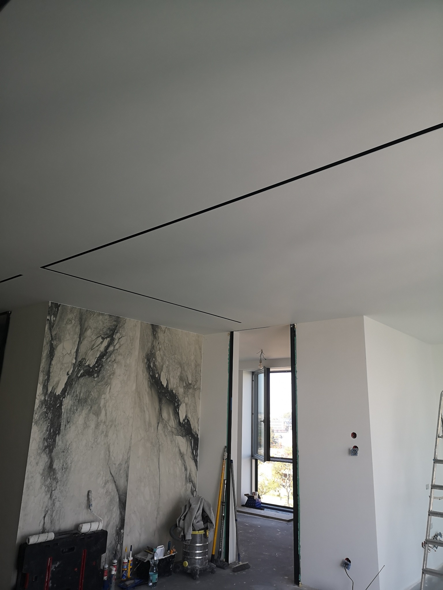 Wnętrze w trakcie prac wykończeniowych: biały sufit z czarnymi listwami, ściana z efektem marmuru, widoczne narzędzia malarskie i drabina. Okno z widokiem na zewnątrz.