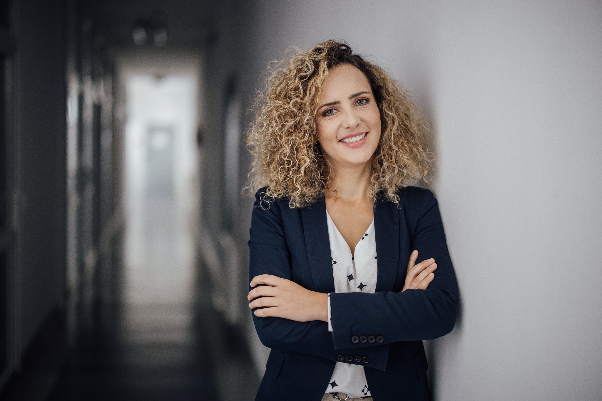 Uśmiechnięta prawniczka w granatowej marynarce, oparta o ścianę na rozmytym tle korytarza. Profesjonalny wizerunek, budzący zaufanie.