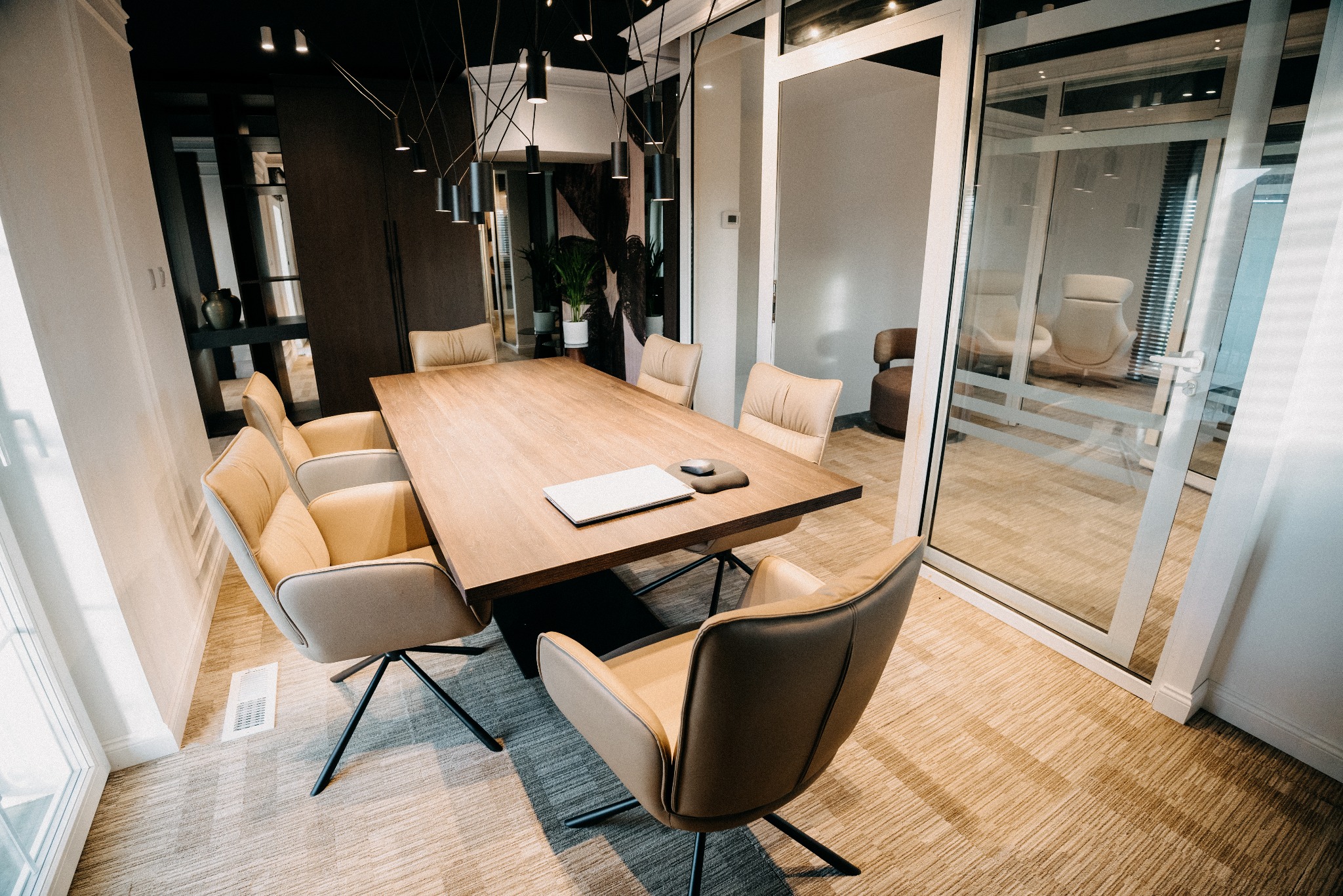 Nowoczesna sala konferencyjna z drewnianym stołem, krzesłami w odcieniach beżu i szklaną ścianą. Na stole leży laptop i myszka. Minimalistyczny design, stonowana kolorystyka.