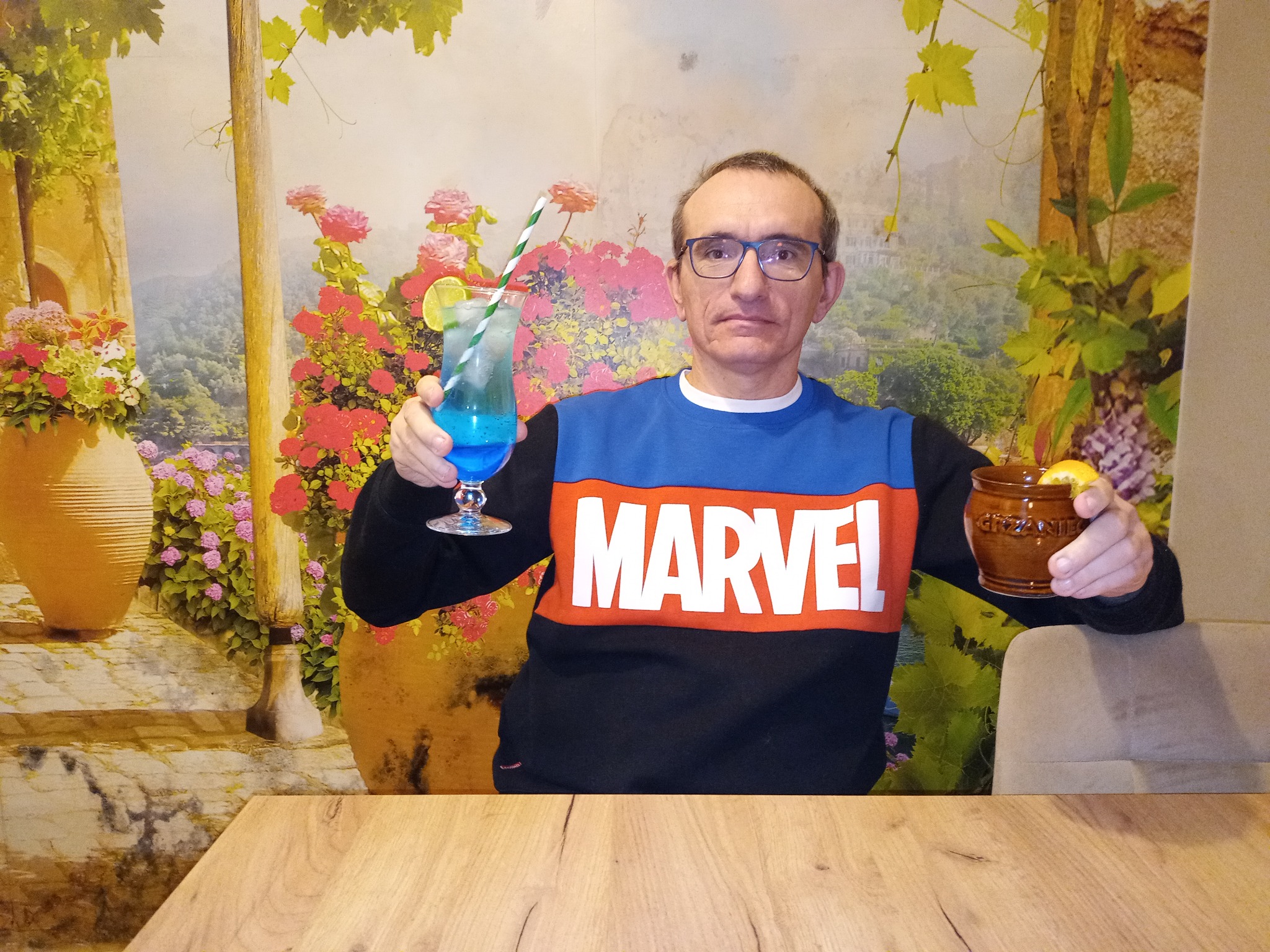 Mężczyzna w bluzie Marvel trzyma koktajl z limonką i napój w glinianym naczyniu na tle ściany z motywem kwiatowym.