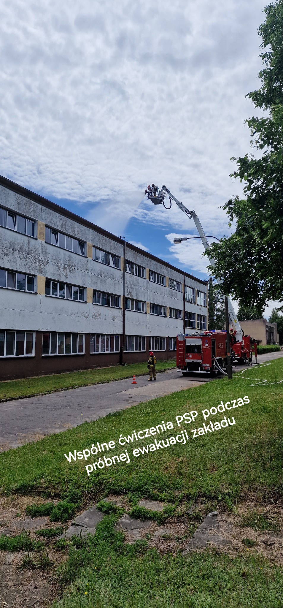 Ćwiczenia PSP: wóz strażacki z wysięgnikiem przy budynku, strażak w hełmie i mundurze, stożek ostrzegawczy. Tekst: Wspólne ćwiczenia PSP podczas próbnej ewakuacji zakładu.