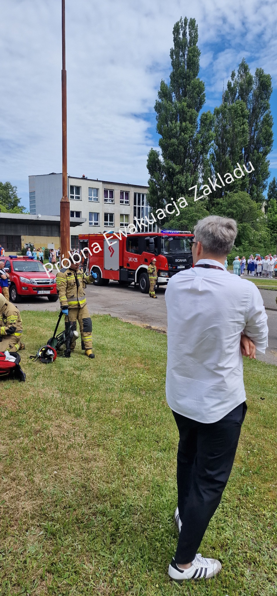 Próbna ewakuacja zakładu: strażacy w akcji, w tle budynek i zgromadzeni ludzie. W kadrze wóz strażacki Scania i czerwony samochód osobowy. Widok z perspektywy obserwatora.