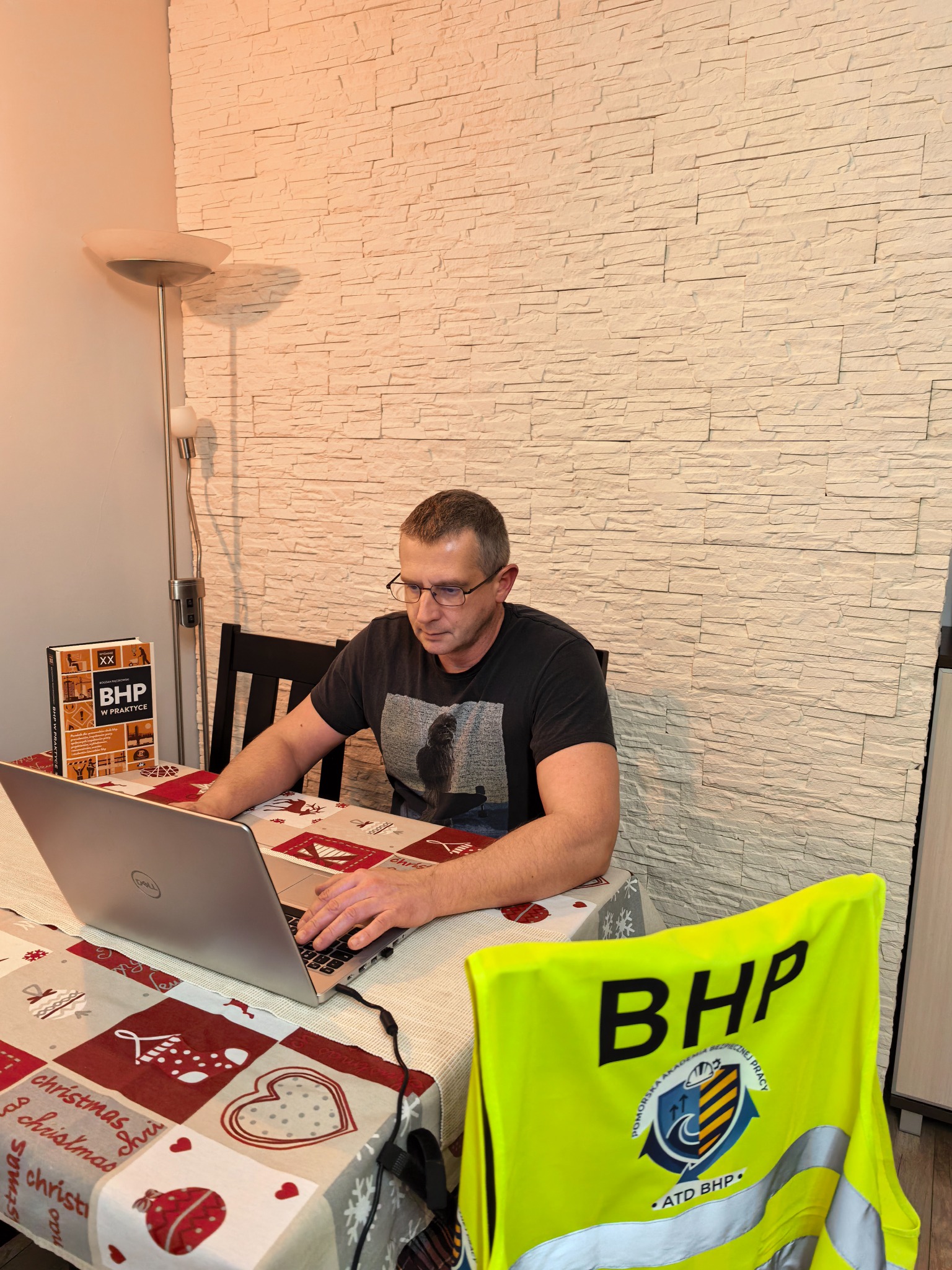 Mężczyzna w okularach pracuje na laptopie przy stole z książką BHP i kamizelką odblaskową BHP, na tle ściany z dekoracyjnej cegły.