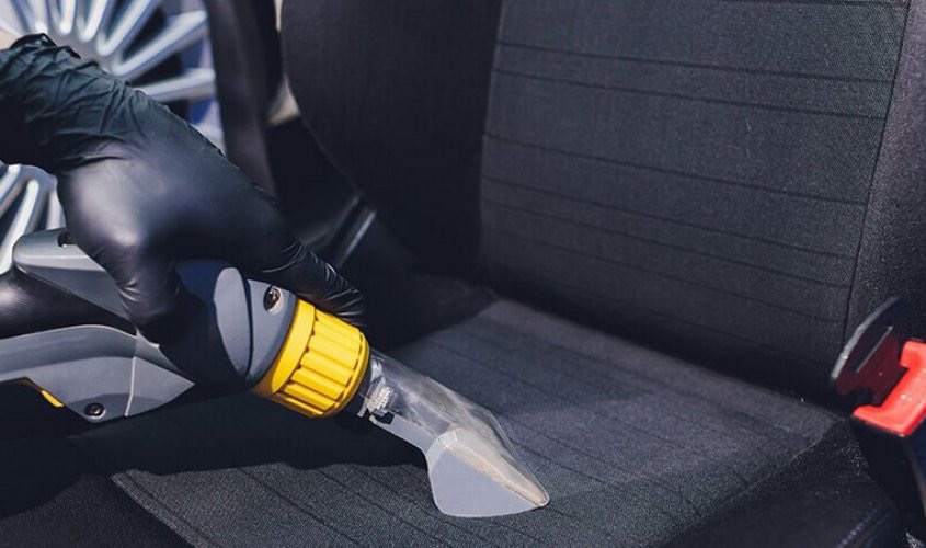 Czyszczenie ciemnej tapicerki samochodowej parownicą. Dłoń w czarnej rękawicy obsługuje urządzenie z żółtymi elementami. Widoczne fragmenty fotela i zagłówka.