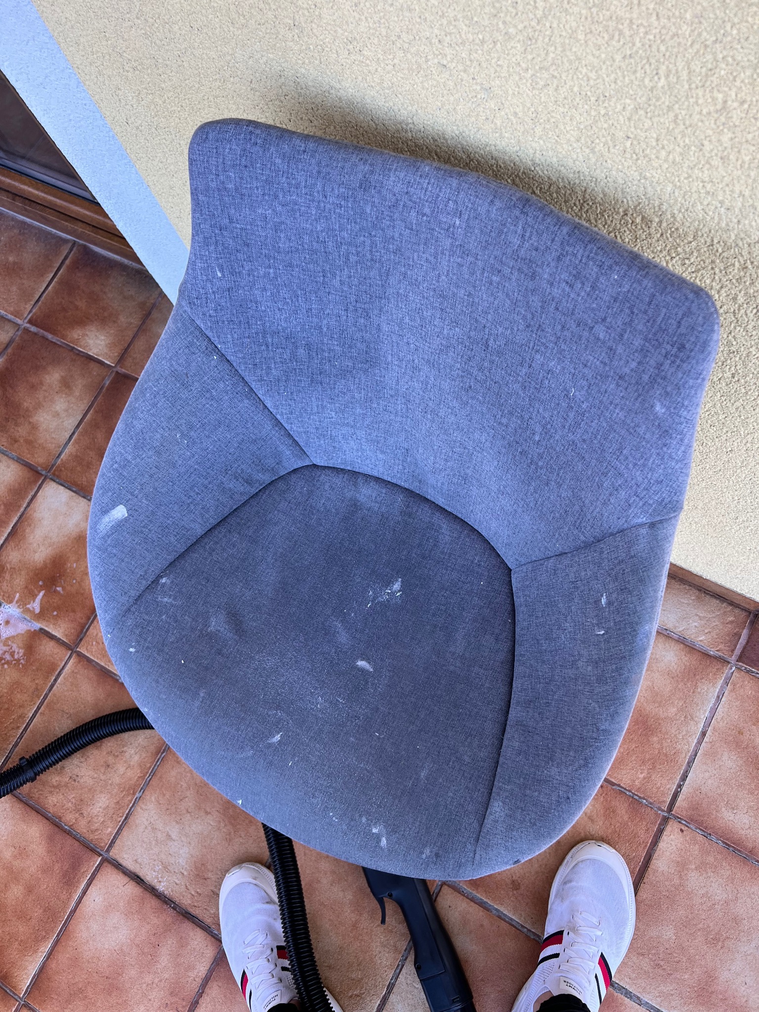 Szare krzesło tapicerowane z plamami, częściowo wyczyszczone, leży na brązowych płytkach. Widoczny wąż odkurzacza i fragment buta osoby czyszczącej.