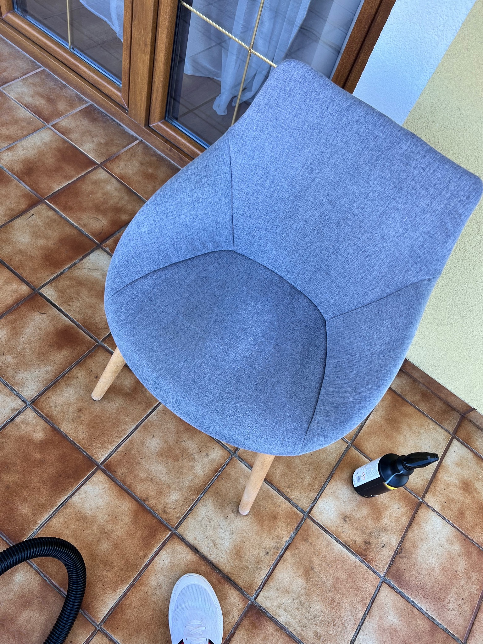 Szare krzesło tapicerowane na brązowych płytkach, obok butelka środka czyszczącego i wąż odkurzacza. Widoczny fragment buta. Ujęcie z góry.