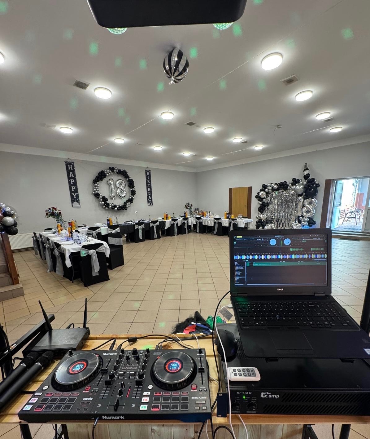 Sala balowa z dekoracją na 18 urodziny, widok z perspektywy DJ-a. Sprzęt DJ-ski na pierwszym planie, w tle stoły z czarno-białymi dekoracjami i balonami.