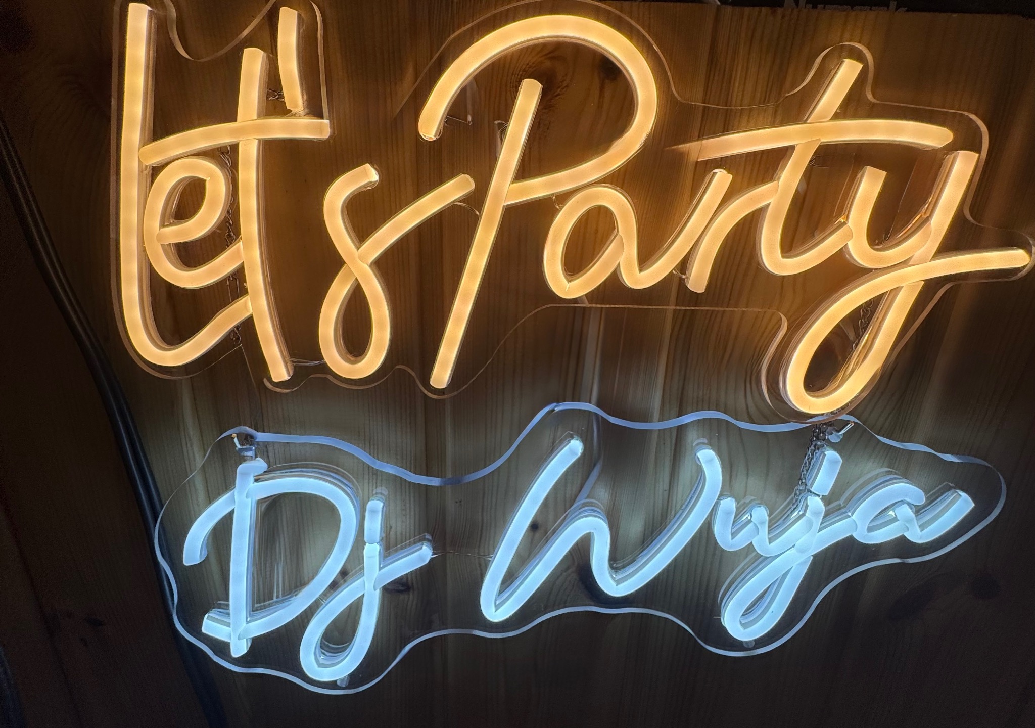 Dwa neony: górny złoty z napisem 'Let's Party', dolny biały z napisem 'DJ Wuja', zawieszone na drewnianej ścianie. Dekoracja świetlna na imprezę.