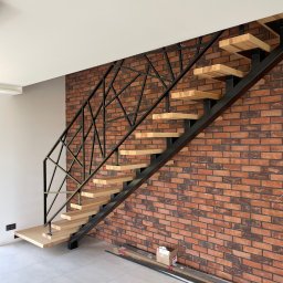 IronBox - Nowoczesne schody metalowe z drewnianymi stopniami, czarna, geometryczna balustrada na tle ceglanej ściany. Widoczne elementy montażowe na podłodze.