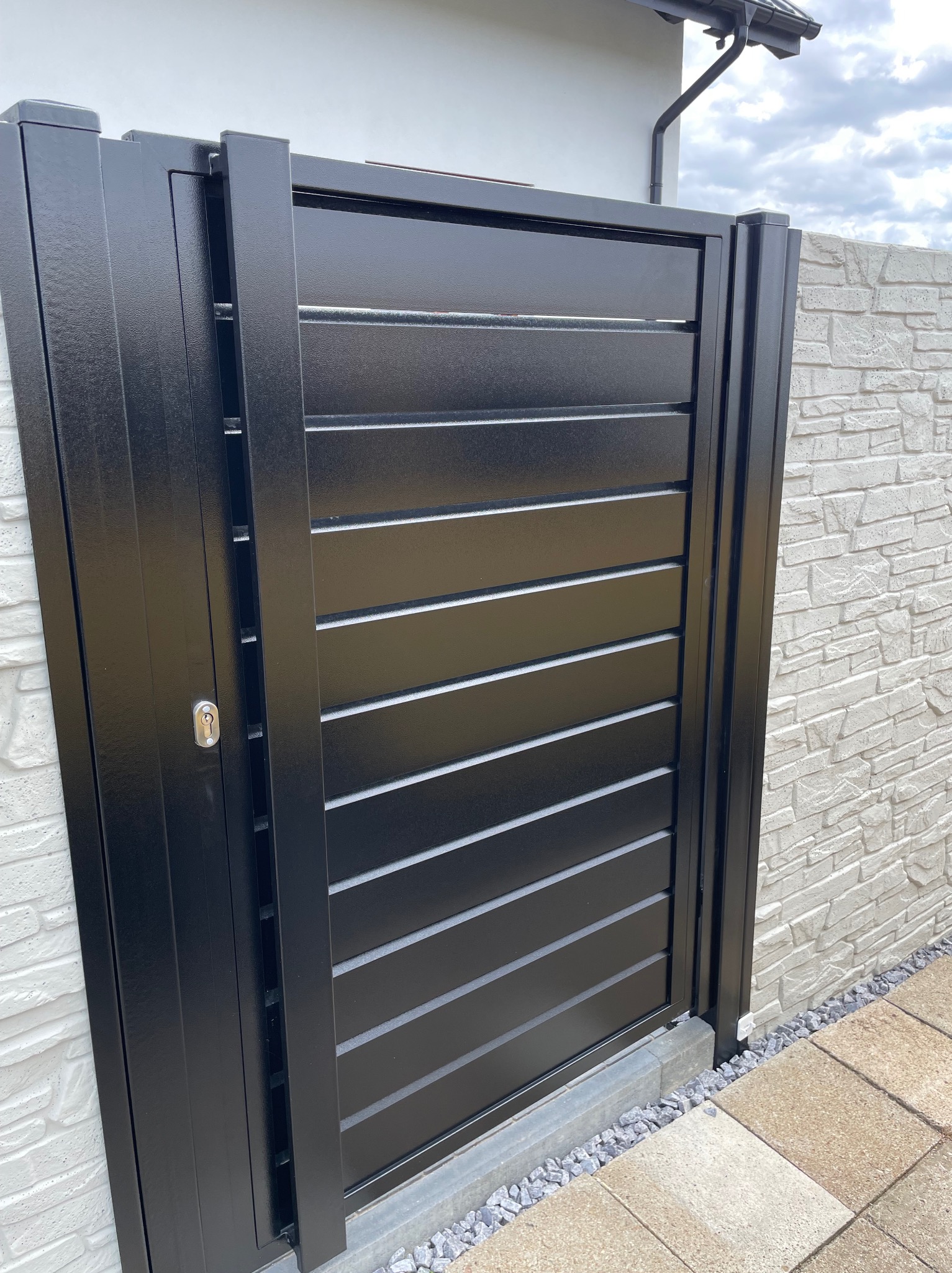 Nowoczesna, czarna brama wjazdowa z poziomymi panelami, zamontowana przy białym, teksturowanym murze. Widoczny zamek i fragment chodnika z kostki brukowej.