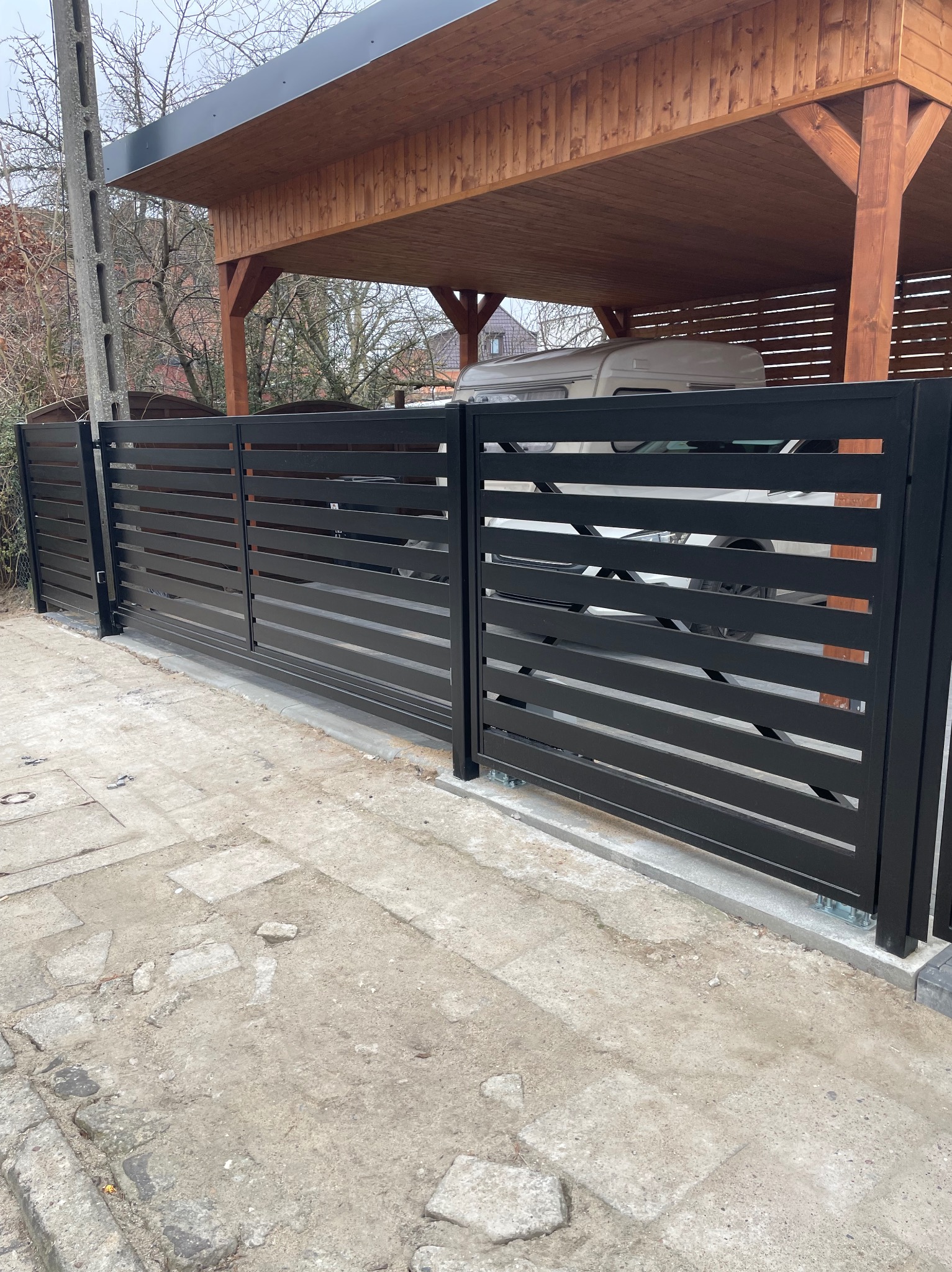 Nowoczesna, przesuwna brama metalowa z poziomymi szczeblami w kolorze czarnym, zamontowana przed drewnianą wiatą garażową. Widoczne betonowe podłoże i słup energetyczny.