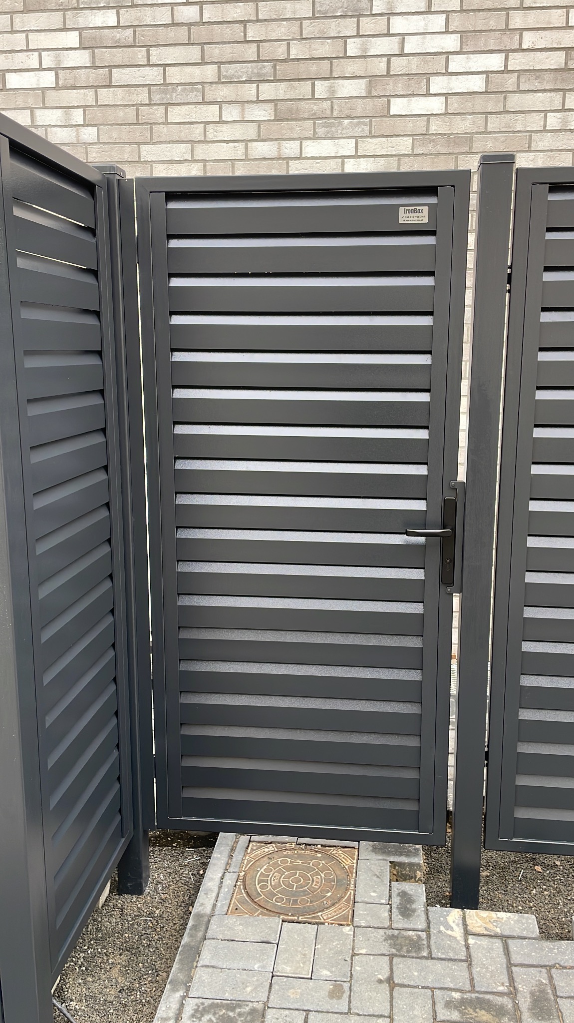 Nowoczesna, grafitowa brama wjazdowa z poziomymi lamelami, zamontowana przy ogrodzeniu z tego samego materiału. Widoczny fragment muru z cegły w tle.