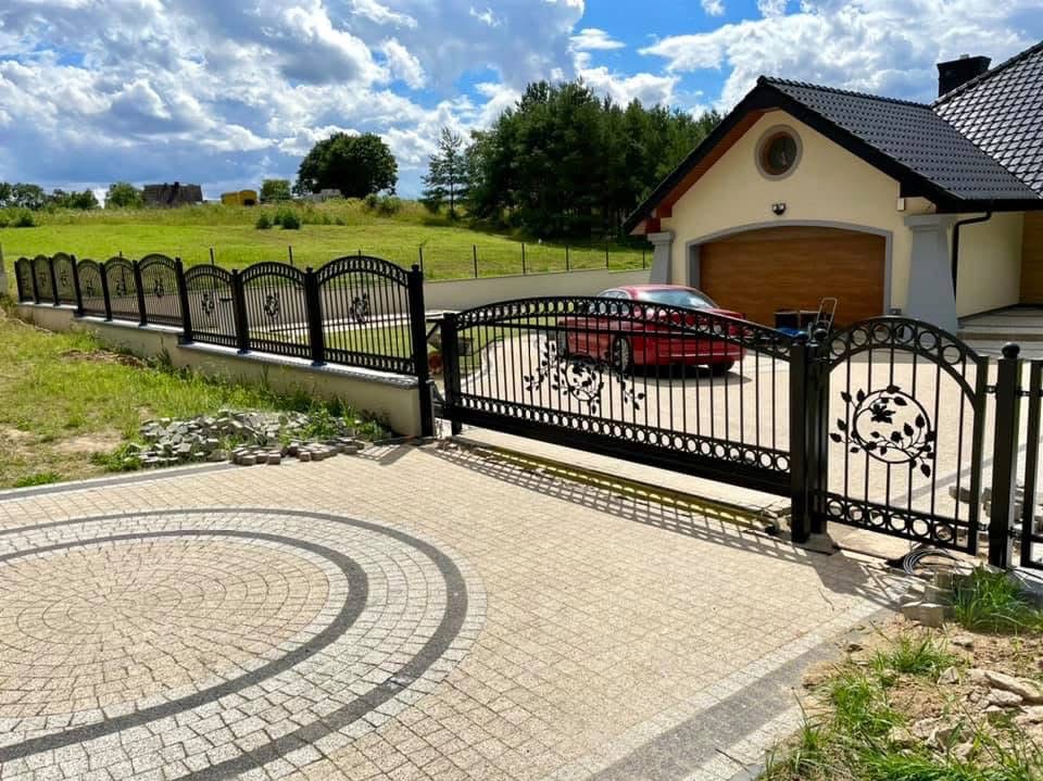 Czarna, metalowa brama wjazdowa z dekoracyjnymi elementami roślinnymi, dopasowana do ogrodzenia na tle domu z garażem i brukowanym podjazdem.
