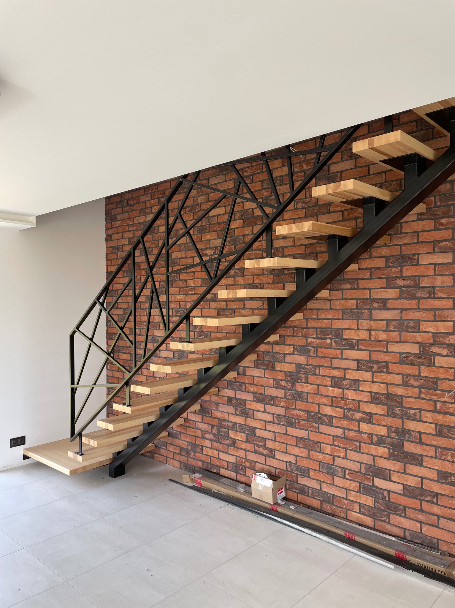 Nowoczesne schody metalowe z drewnianymi stopniami, czarna, geometryczna balustrada na tle ceglanej ściany. Widoczne elementy montażowe na podłodze.
