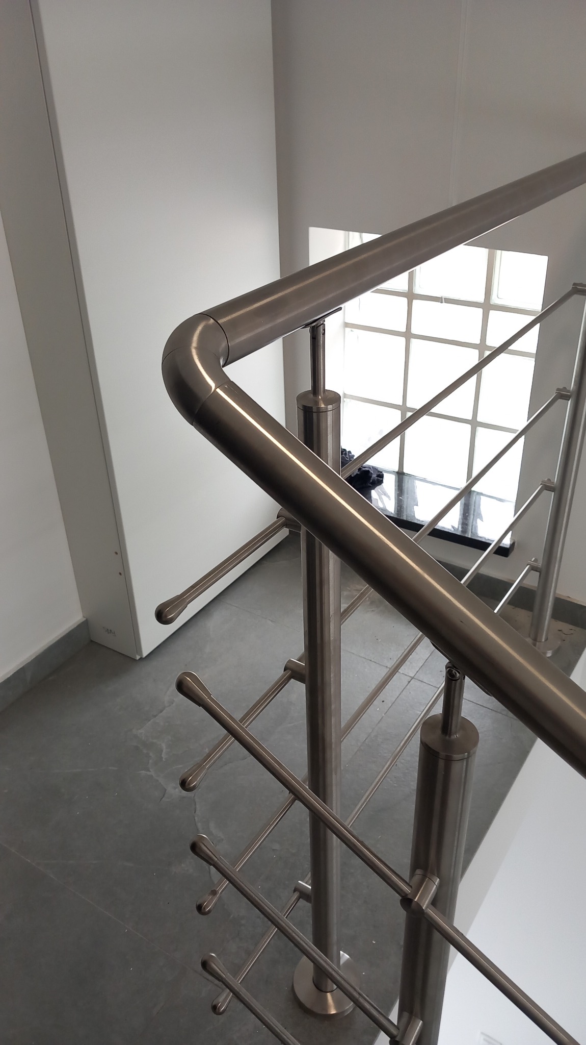 Nowoczesna balustrada schodowa ze stali nierdzewnej, widok z góry. Minimalistyczny design z poziomymi prętami i zaokrąglonym poręczą. Jasne wnętrze z oknem w tle.