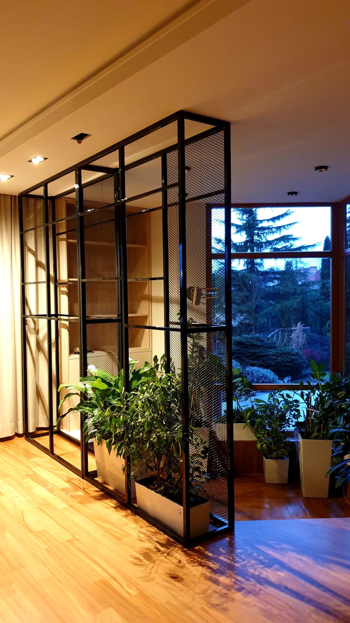 Loftowa ścianka działowa z metalową siatką, dzieląca przestrzeń w jasnym wnętrzu. Widoczne rośliny doniczkowe i okno z widokiem na zieleń.