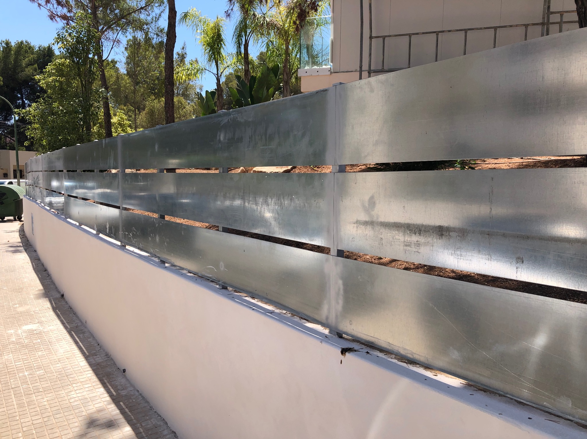 Nowoczesne ogrodzenie metalowe z poziomych paneli, z widocznymi przerwami między nimi. Biały murek stanowi podstawę ogrodzenia. W tle zieleń drzew i fragment budynku.